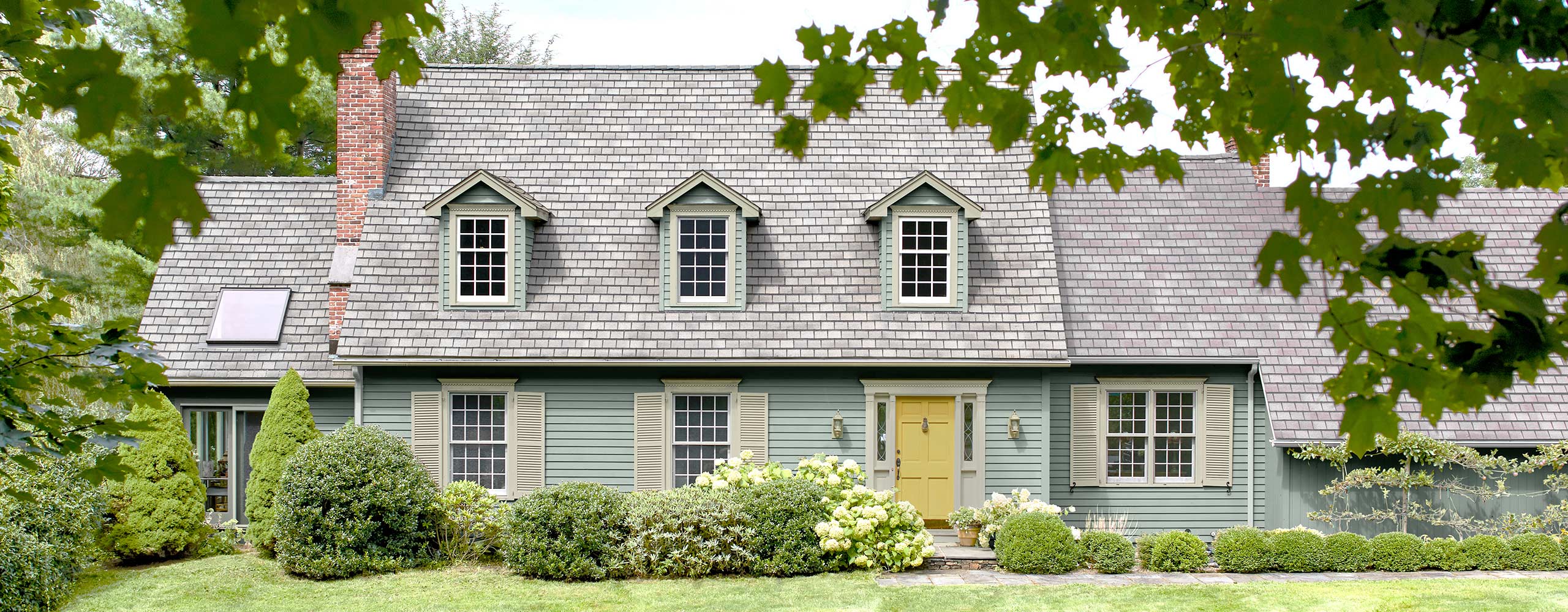 A beautiful cape style home with soft blue-gray painted siding, pale green shutters and trim, a yellow door, dormer windows, a lush green lawn and shrubbery. A beautiful cape style home with soft blue-gray painted siding, pale green shutters and trim, a yellow door, dormer windows, a lush green lawn and shrubbery.