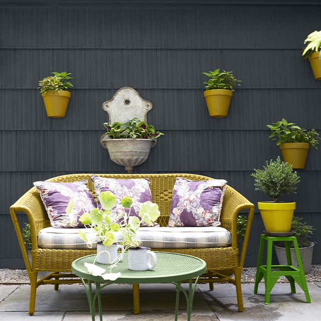 Patio chaleureux avec canapé rembourré en osier jaune-vert et coussins colorés ainsi que deux tables vertes devant un parement noir clair sur lequel sont accrochées des plantes en pot. Patio chaleureux avec canapé rembourré en osier jaune-vert et coussins colorés ainsi que deux tables vertes devant un parement noir clair sur lequel sont accrochées des plantes en pot.