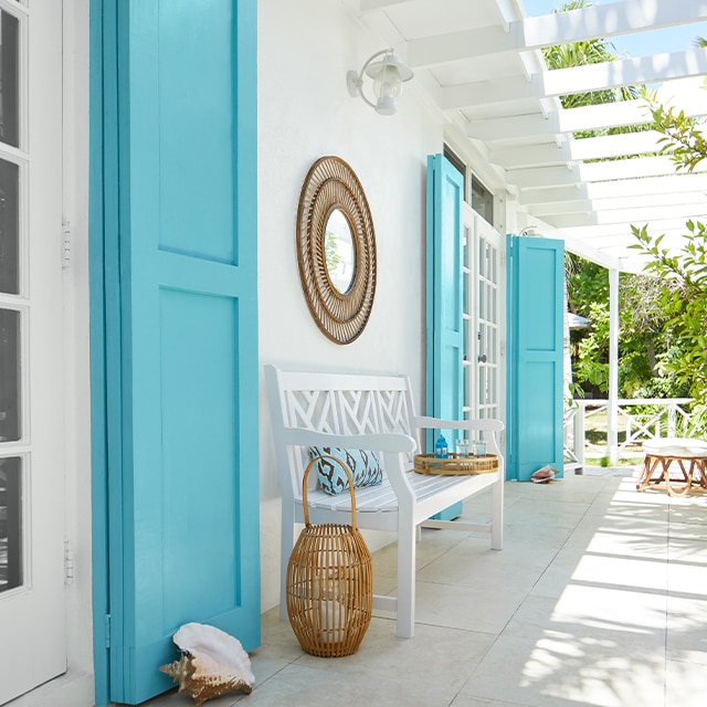 Galerie blanche rappelant les tropiques avec pergola fixée à la maison, longs volets turquoise, banc blanc et accessoires en osier. Galerie blanche rappelant les tropiques avec pergola fixée à la maison, longs volets turquoise, banc blanc et accessoires en osier.