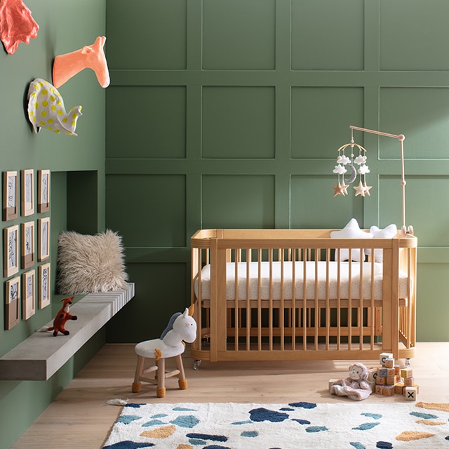 Une chambre de bébé ensoleillée avec un berceau moderne en bois contre un mur en lambris peint en vert riche et un mur vert contigu agrémenté de têtes d’animaux pittoresques suspendues et d’œuvres d’art encadrées, ainsi que d’un tapis multicolore. Une chambre de bébé ensoleillée avec un berceau moderne en bois contre un mur en lambris peint en vert riche et un mur vert contigu agrémenté de têtes d’animaux pittoresques suspendues et d’œuvres d’art encadrées, ainsi que d’un tapis multicolore.