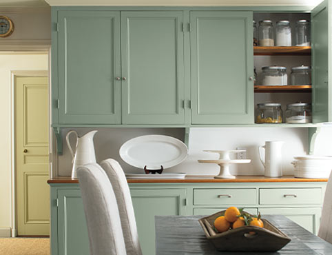 Light green-painted cabinets and white dinnerware add a soft glow as a backdrop to a dining table and white slipcovered chairs.
