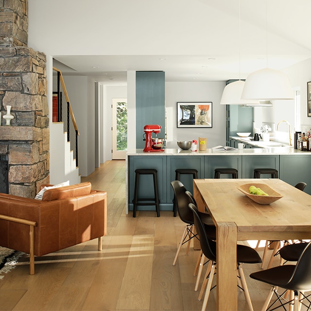 An open floor plan living space with white-painted walls and vaulted ceiling, dark gray kitchen cabinets, a wooden dining table and black chairs, and stone fireplace. An open floor plan living space with white-painted walls and vaulted ceiling, dark gray kitchen cabinets, a wooden dining table and black chairs, and stone fireplace.