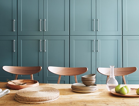 Mur d’armoires bleu sarcelle clair devant lequel se trouve une table en bois avec des napperons en osier, de la vaissellerie en bois et trois chaises en bois. Mur d’armoires bleu sarcelle clair devant lequel se trouve une table en bois avec des napperons en osier, de la vaissellerie en bois et trois chaises en bois.