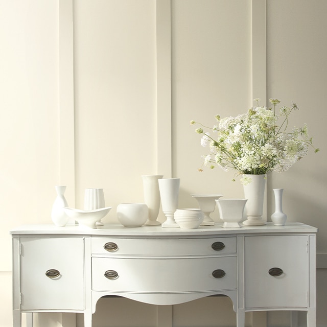 An off-white wall with floor-to-ceiling wainscotting and multiple white vases placed on top of a white dresser.