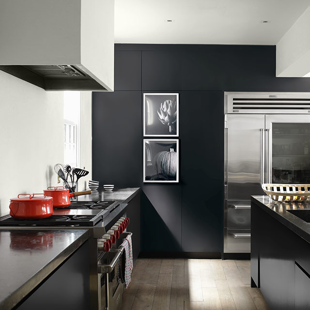 A sleek, contemporary kitchen with white-painted walls and ceiling, a black accent wall, painted black cabinets, and a large island, open to a white hallway. A sleek, contemporary kitchen with white-painted walls and ceiling, a black accent wall, painted black cabinets, and a large island, open to a white hallway.