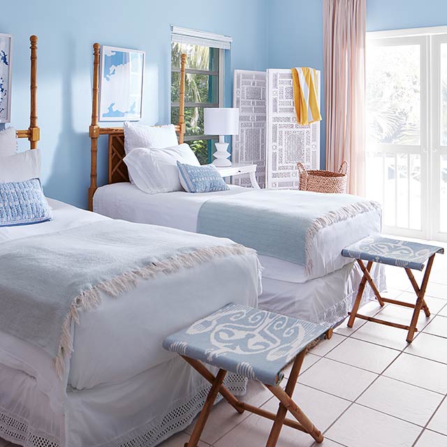 Chambre aux murs bleus avec moulures et plafond blancs, lits jumeaux et tabourets pliants dans un décor bleu et blanc.