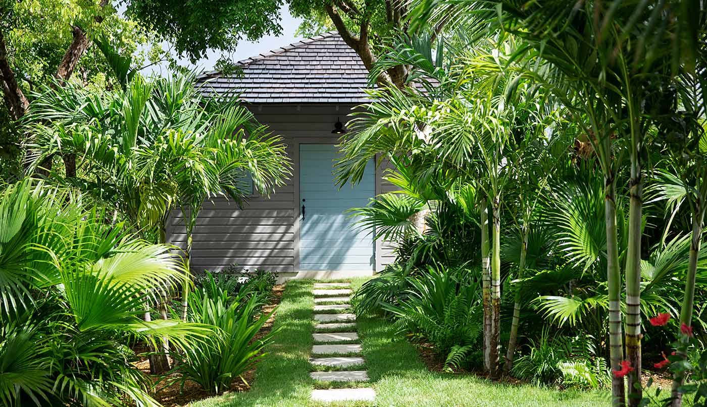 Maison peinte en gris avec porte bleue et chemin d’accès en pierre dans un décor d’arbres et de végétation tropicaux.