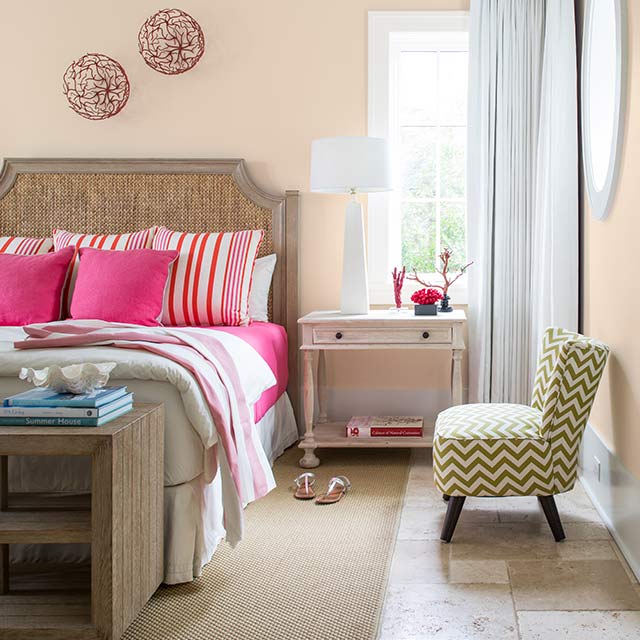Peach-painted bedroom walls with a green chevron chair, side table, and bed featuring pink bedding.