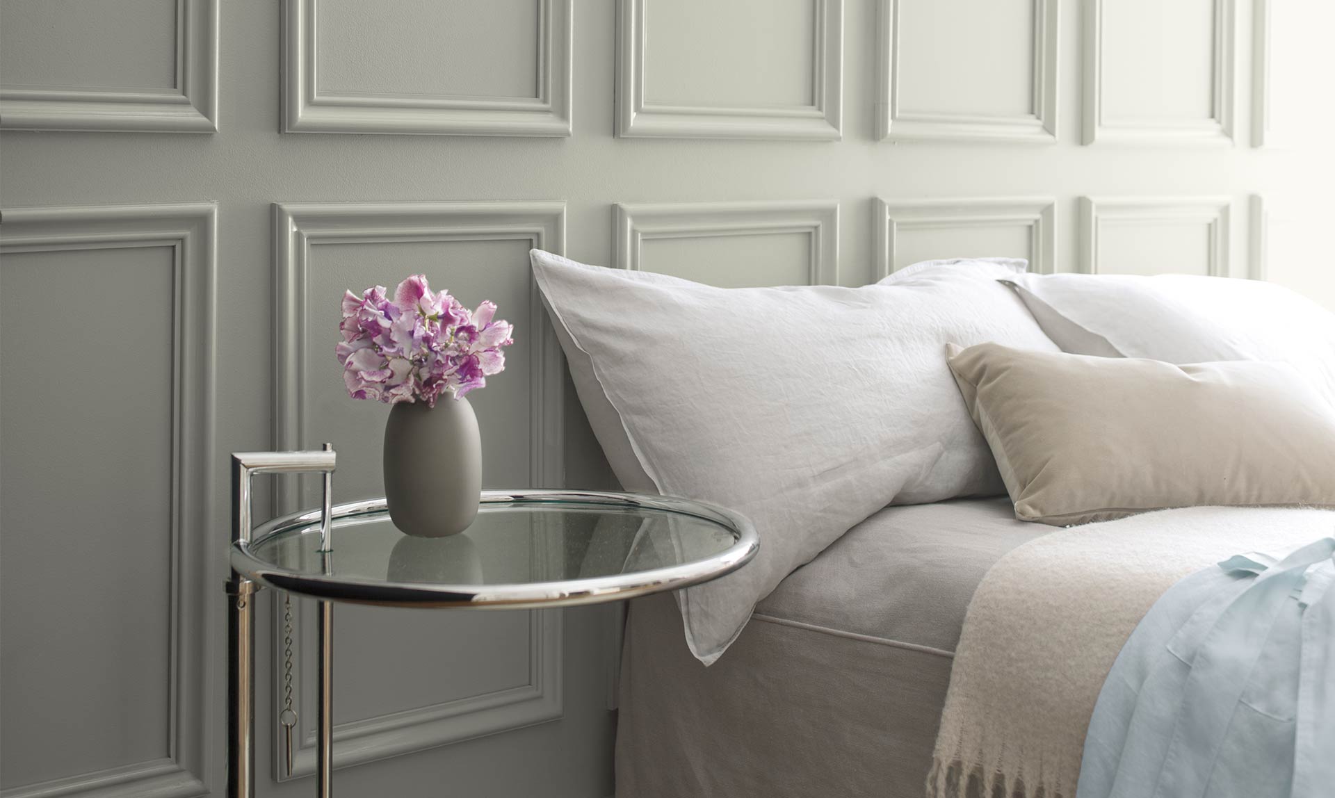Un lit à la literie neutre est placé contre un mur gris et à côté d’une table de nuit en verre. Couleur de l’année 2019 Un lit à la literie neutre est placé contre un mur gris et à côté d’une table de nuit en verre. Couleur de l’année 2019